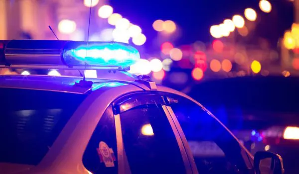 A police car with flashing blue lights is parked at night with blurred city lights in the background.