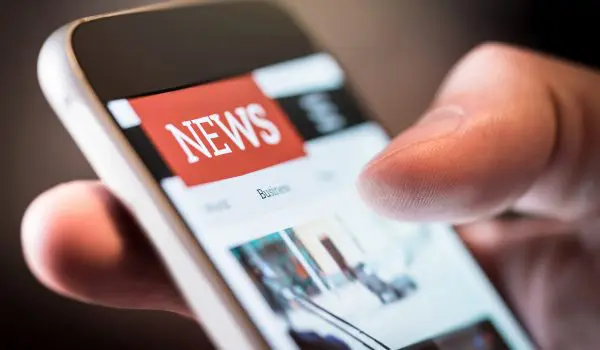 A close-up view of a person's hand holding a smartphone displaying a news website with a red "NEWS" banner at the top.