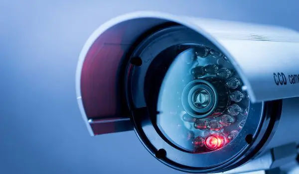 A close-up of a security camera with a red recording light, mounted on a wall against a blue background.