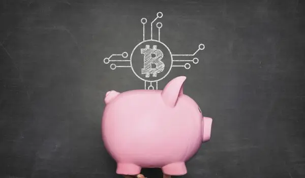 A hand holds a pink piggy bank in front of a blackboard with a Bitcoin symbol and circuit lines drawn on it.