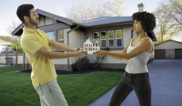 Two people are pulling on a small model house while standing in front of a suburban home.