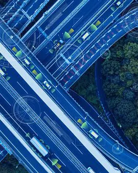 An aerial view of a complex highway interchange with multiple layers of roads and vehicles next to a forested area.