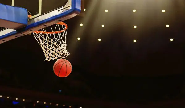 A basketball is mid-air heading towards a hoop during a game under bright lights.