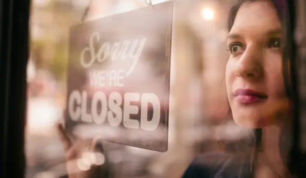 A person is standing inside, looking out a window with a "Sorry, we're closed" sign hanging in front.