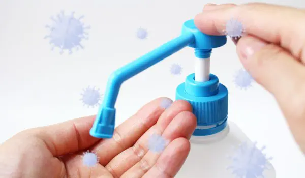 A person is using a hand sanitizer dispenser while surrounded by illustrated virus particles.