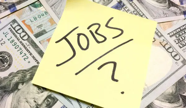 A sticky note with the word "JOBS" written on it is placed on top of a pile of U.S. hundred-dollar bills.