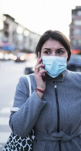 A person wearing a face mask and carrying a patterned bag is talking on the phone while standing at a crosswalk on a city street.