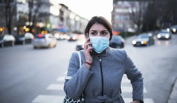 A person wearing a face mask and carrying a patterned bag is talking on the phone while standing at a crosswalk on a city street.