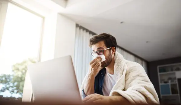 A person wrapped in a blanket works on a laptop while holding a tissue to their nose.