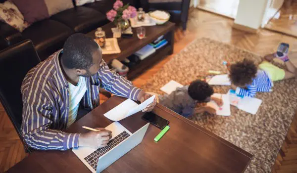 A person works on a laptop at a table while two children draw on the floor nearby.