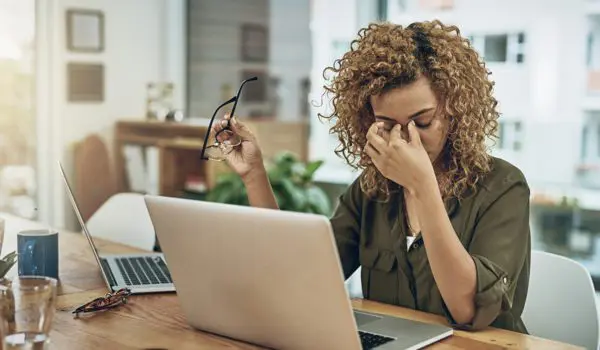 A person sitting at a desk with a laptop, holding their glasses and rubbing their eyes.