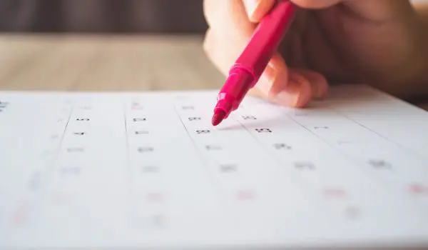 A hand is using a red pen to mark a date on a calendar.