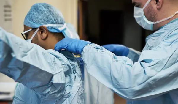 Two medical professionals wearing protective gear are assisting each other with their gowns.