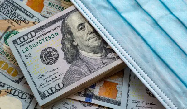 A stack of hundred-dollar bills is partially covered by a blue surgical mask.