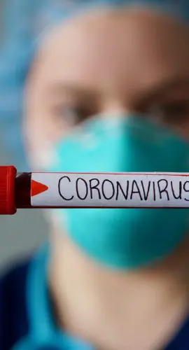 A healthcare worker wearing protective gear holds up a test tube labeled "CORONAVIRUS."