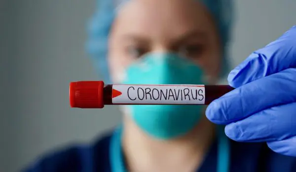A healthcare worker wearing protective gear holds up a test tube labeled "CORONAVIRUS."