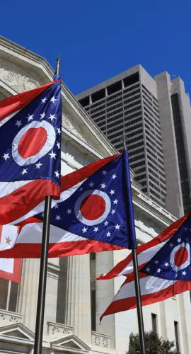 A series of Ohio state flags are flying in front of a classical building under a clear blue sky, with tall modern buildings in the background.