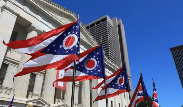 A series of Ohio state flags are flying in front of a classical building under a clear blue sky, with tall modern buildings in the background.
