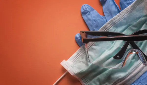 A pair of eyeglasses sits on top of a face mask and blue medical gloves against an orange background.