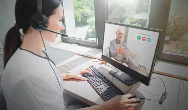 A woman wearing a headset is using a computer to have a video call with an older man who appears on the screen.