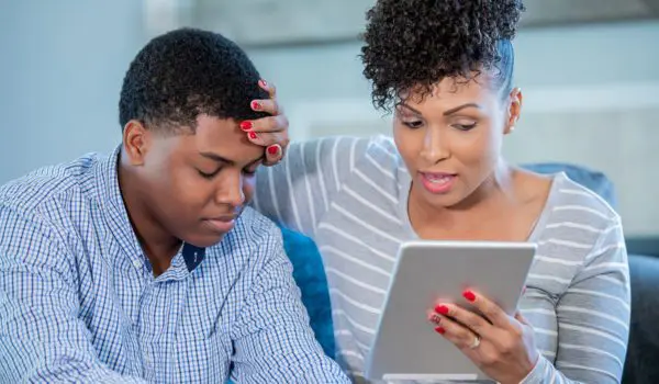 A woman is looking at a tablet while placing her hand on the forehead of a person sitting next to her.