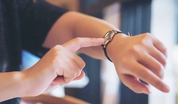 A person is pointing at their wristwatch while wearing a dark sleeve.