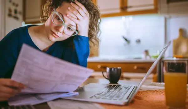 A person with curly hair and glasses appears stressed while looking at documents next to a laptop, a cup, and a glass of orange juice in a kitchen setting.