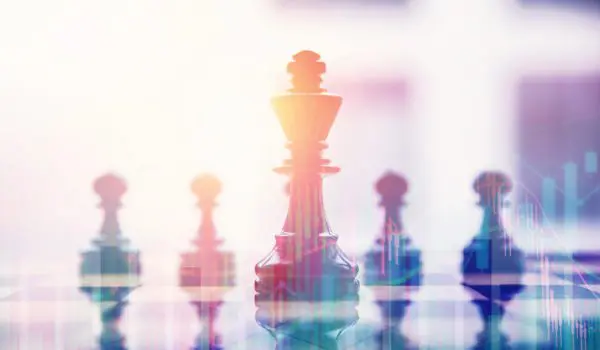 Several chess pieces are arranged on a reflective surface, with a prominent king piece in focus and a bright, abstract background.
