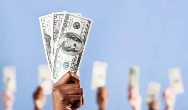 Multiple hands are holding up US dollar bills against a light blue background.