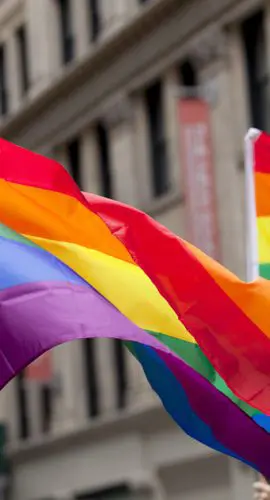 Rainbow flags are waving in front of a series of beige buildings.