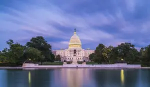 The U.S. Capitol building is lit up against a twilight sky with a reflection on a calm body of water in the foreground.