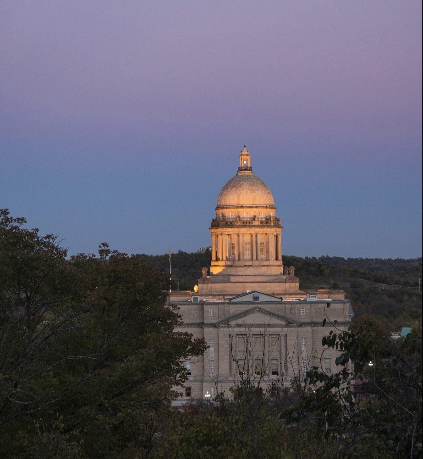 A grand building with a prominent dome is illuminated against a twilight sky, surrounded by trees.