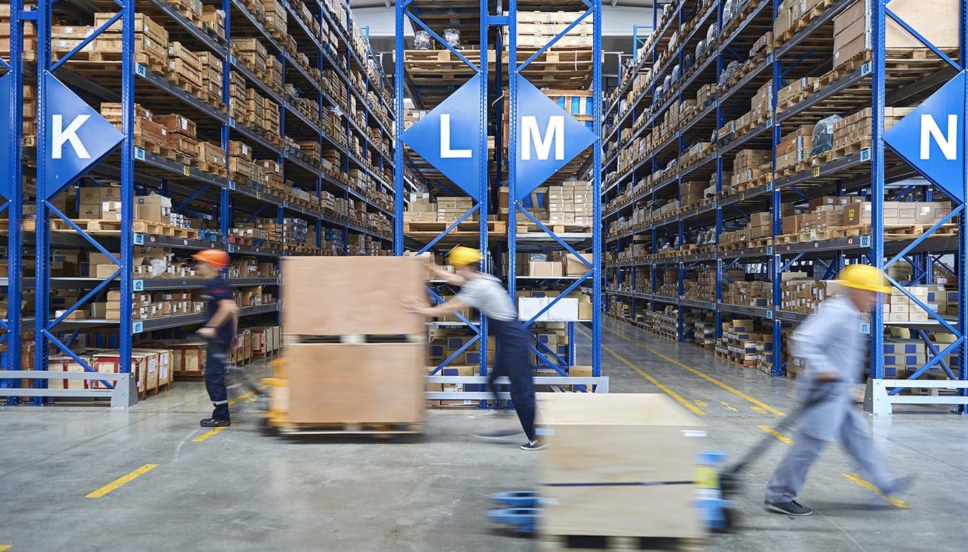 Workers in hard hats are actively moving goods around in a well-organized warehouse filled with tall shelving units.