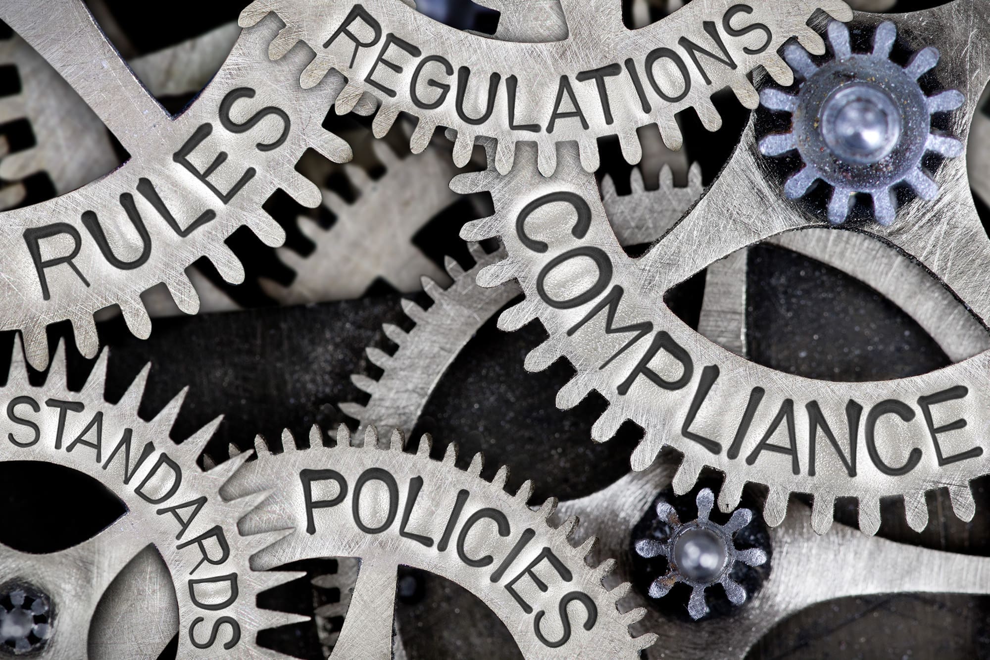 A network of metal gears is inscribed with the words "RULES," "REGULATIONS," "STANDARDS," "POLICIES," and "COMPLIANCE."