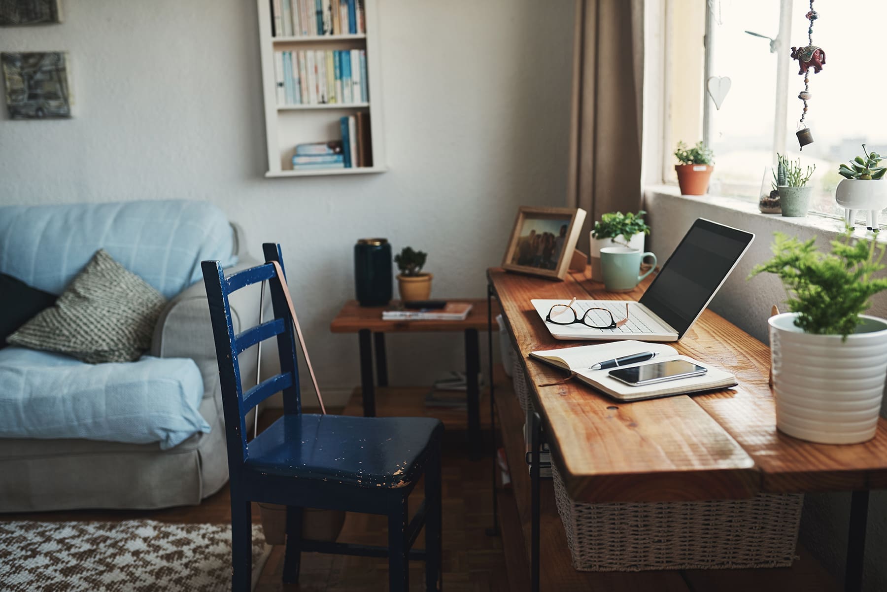 A cozy home office setup with a wooden desk, laptop, books, indoor plants, and a nearby sofa.