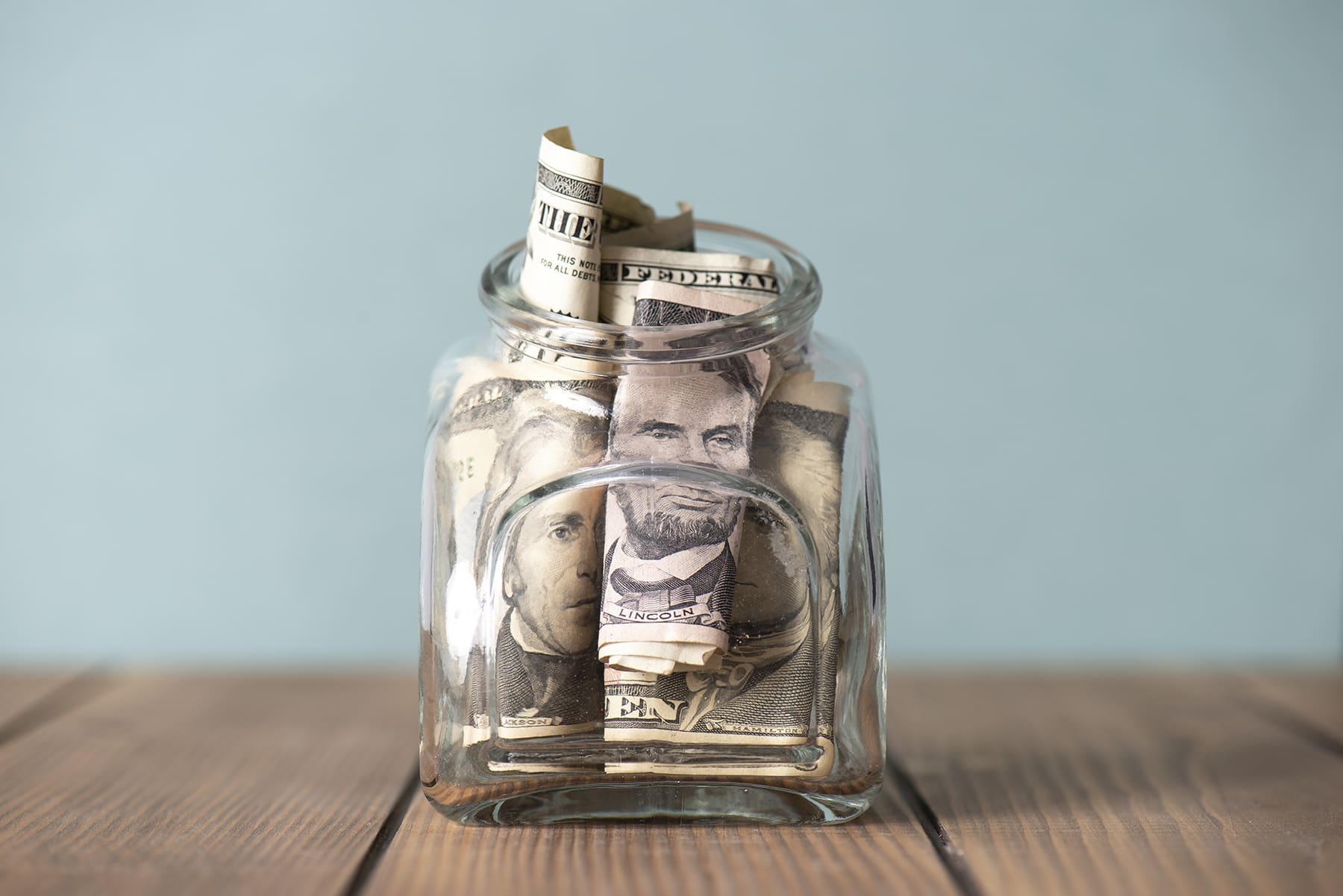 A glass jar filled with rolled and folded U.S. dollar bills sits on a wooden surface against a plain, light-colored background.