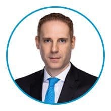 A man with short, reddish-brown hair is wearing a black suit, white shirt, and blue tie, framed within a blue circle against a white background.