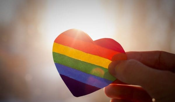 hand holds a heart painted like a LGBT flag, silhouetted against sun