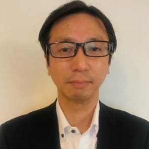A person with short black hair and glasses is wearing a white shirt and black blazer against a plain background.