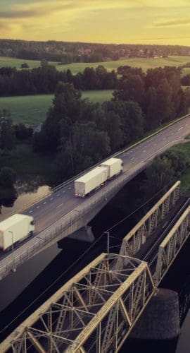 Two trucks with trailers on the road at sunset through a rural landscape in the Dalarna region of Sweden.