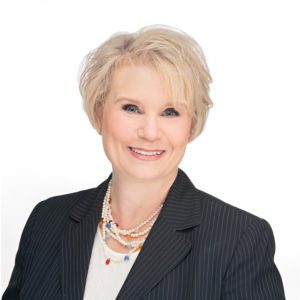 A person with short, light-colored hair is wearing a dark pinstripe suit and layered necklaces, smiling against a white background.