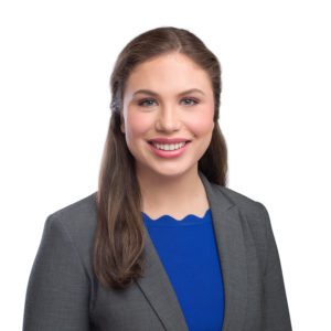 A smiling person with long brown hair is dressed in a gray blazer over a blue top in front of a white background.