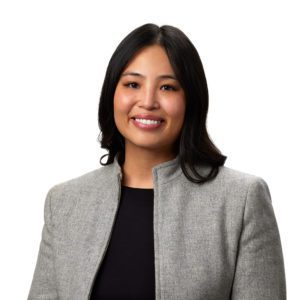 A person wearing a grey jacket over a black top stands against a plain white background, smiling.