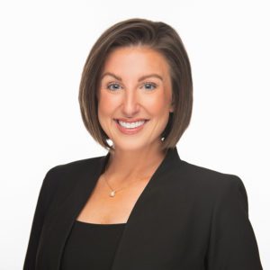 A person is smiling, dressed in a black top and blazer, with short brown hair against a white background.