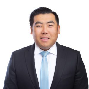 A person wearing a black suit, white shirt, and light blue tie is smiling against a white background.