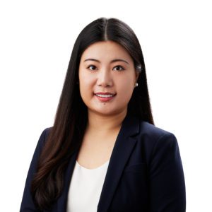 A person with long dark hair is wearing a navy blazer and white shirt, smiling in front of a plain background.