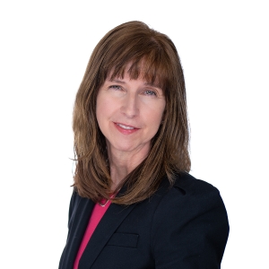 A person with shoulder-length brown hair is wearing a black blazer over a red top, posing against a plain white background.