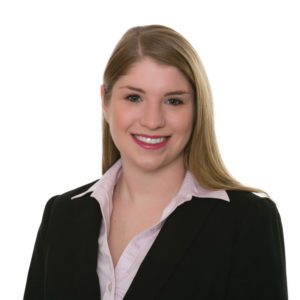 A person with straight blonde hair, wearing a black blazer and a white collared shirt, is smiling against a white background.