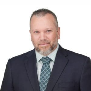 A man with short, graying hair and a beard, dressed in a suit and tie, poses against a white background.