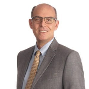 A man wearing a suit, tie, and glasses is smiling against a plain white background.
